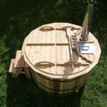 Round wooden barrel hot tub with lid, steps, and a metal chimney on a grassy lawn.